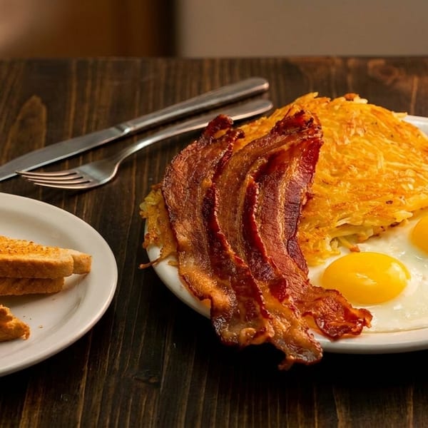 A breakfast plate with crispy bacon, sunny-side-up eggs, hash browns, and toast on a wooden table.