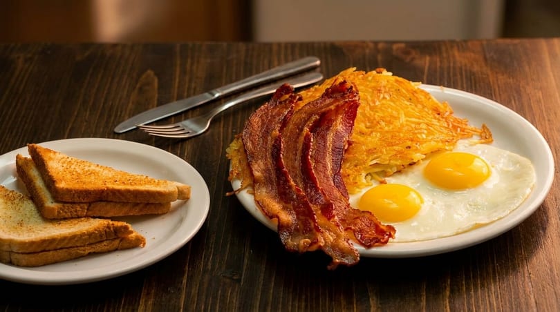 A breakfast plate with crispy bacon, sunny-side-up eggs, hash browns, and toast on a wooden table.