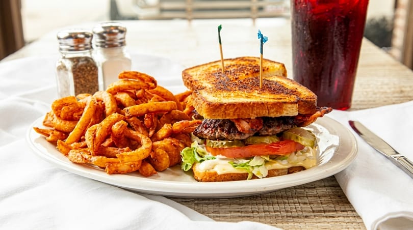 A burger on toasted bread with curly fries, a drink, and salt and pepper shakers on a table.