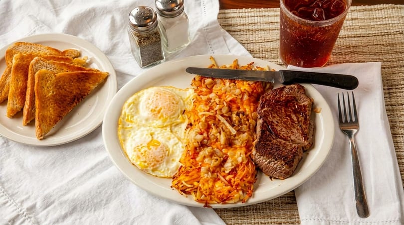 Steak and eggs breakfast with hash browns, toast, and a drink on a white tablecloth.