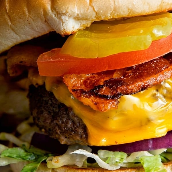 Close-up of a juicy cheeseburger with bacon, tomato, pickles, red onion, and lettuce, with fries.