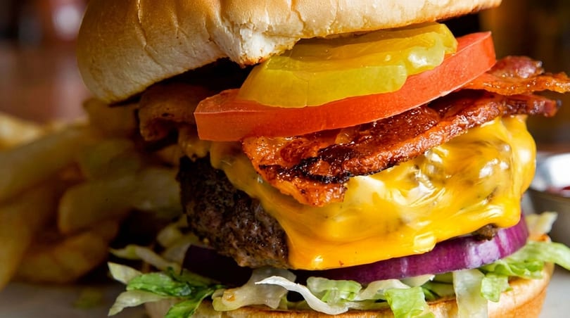 Close-up of a juicy cheeseburger with bacon, tomato, pickles, red onion, and lettuce, with fries.