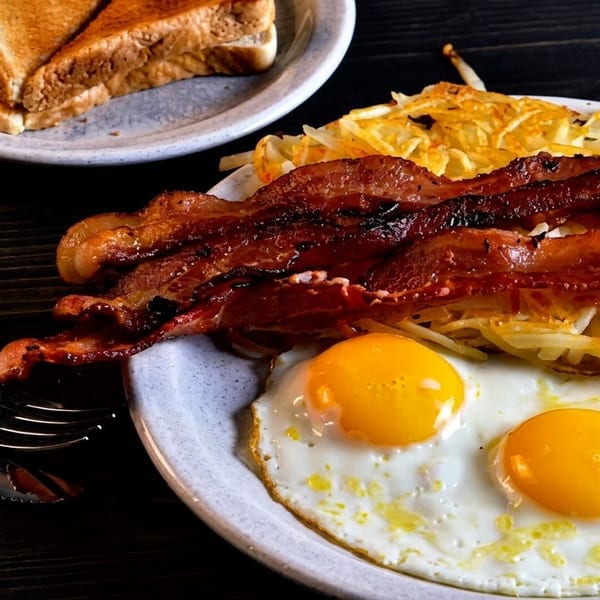 A breakfast plate with crispy bacon, hash browns, two fried eggs, and toast on a dark wooden table.