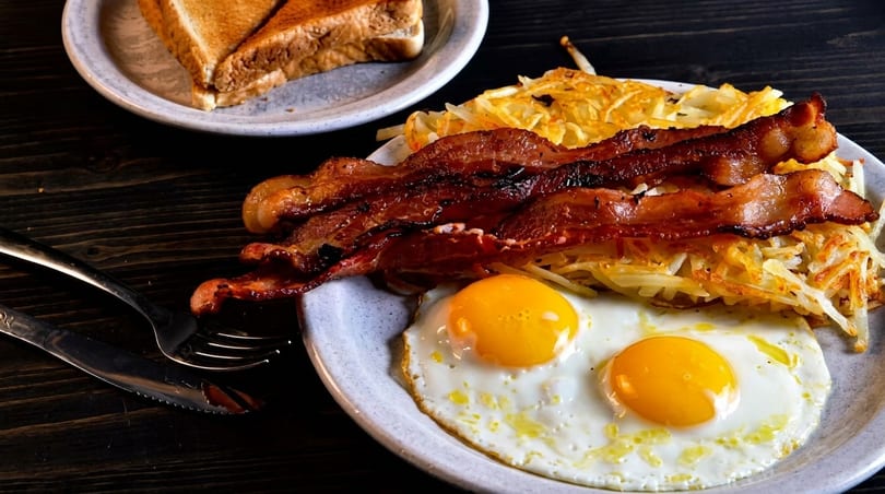 A breakfast plate with crispy bacon, hash browns, two fried eggs, and toast on a dark wooden table.